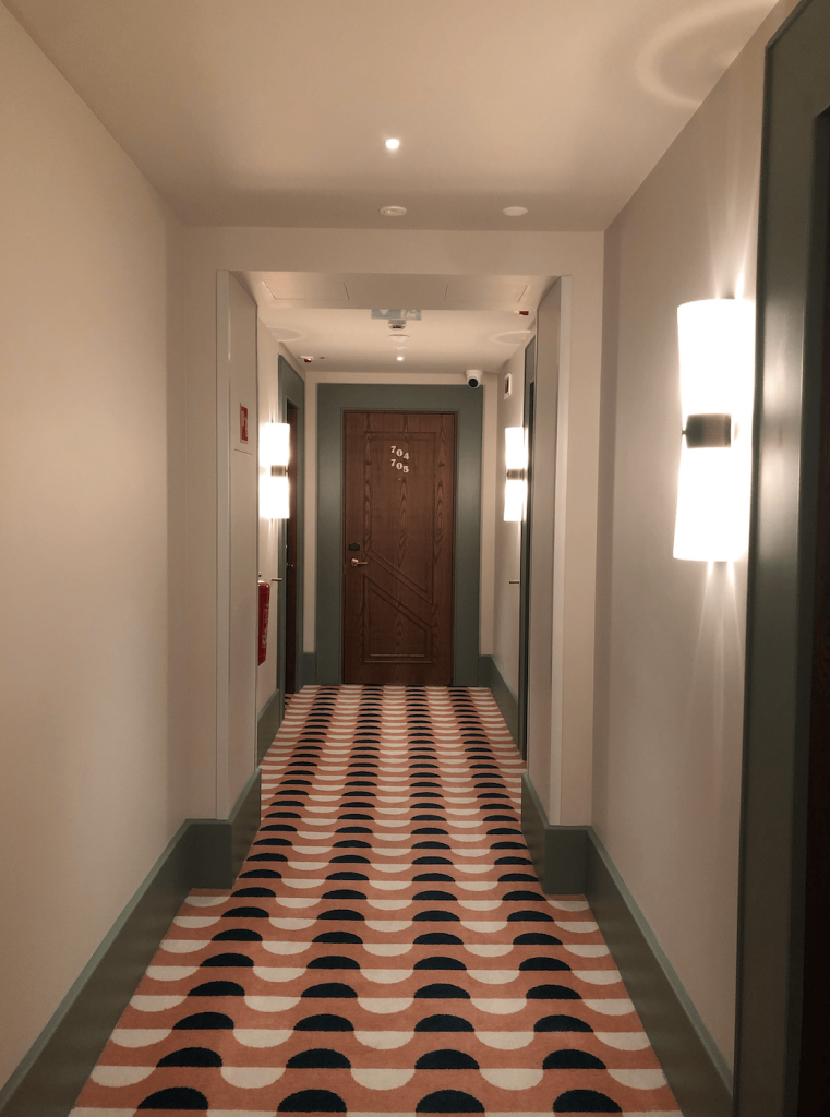 hoxton hotel hallway. A long corridor with geometric carpet, contrast woodwork, and wall lights leading to a dark wood door