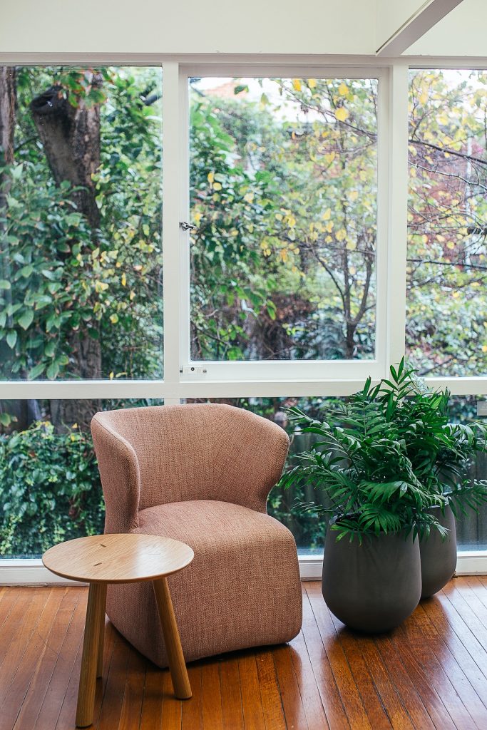 a small upholstered chair in a muted peach colour with wood side table and two fern in pots. Behind are windows looking out on trees and foliage.