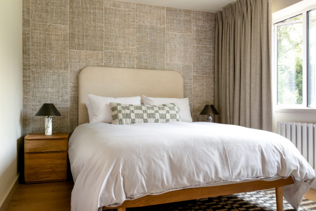 Calm, neutral bedroom with textured wallpaper, natural wood furniture, and floor-length curtains — an example of how soft materials and light tones create an uplifting, airy atmosphere.